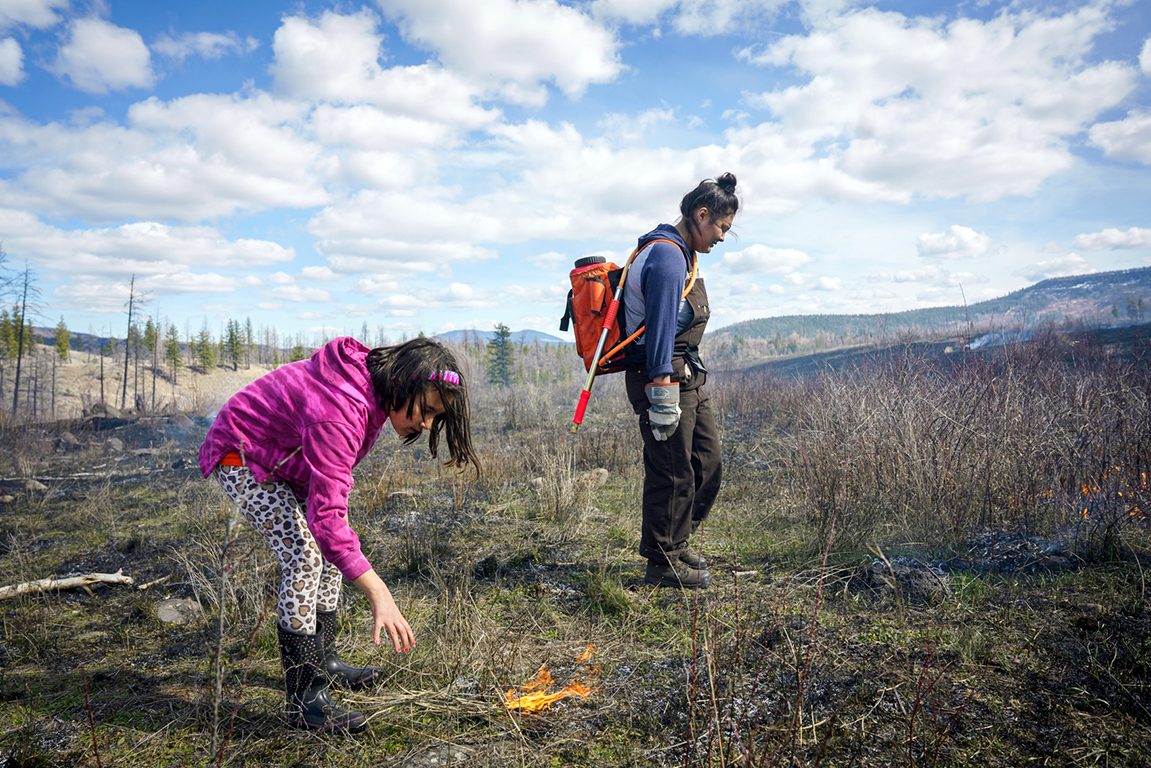 On a ridge with grassy hills behind, in the foreground a girl of about eight years old bends towards the ground where a small flame burns. A woman wearing a water tank on her back strides alongside.