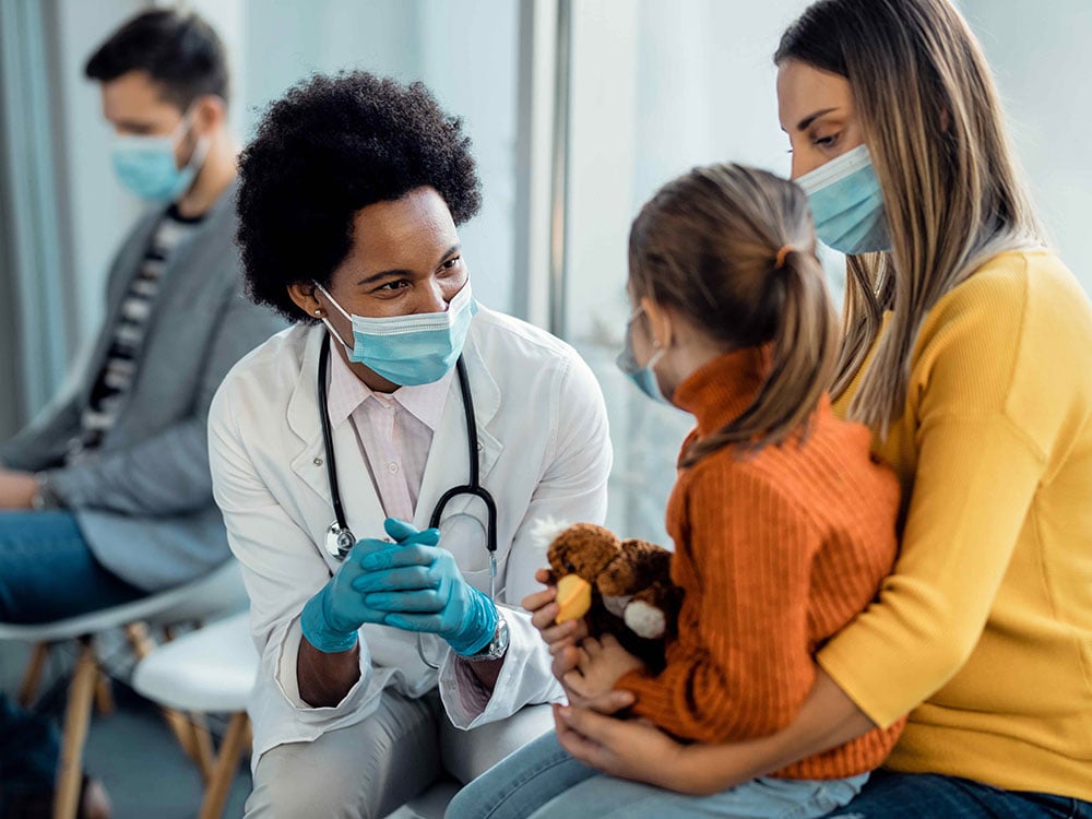 A doctor wearing a white jacket, with stethoscope and face mask, speaks with a young girl who is seated on her mother’s lap holding a stuffed animal toy.