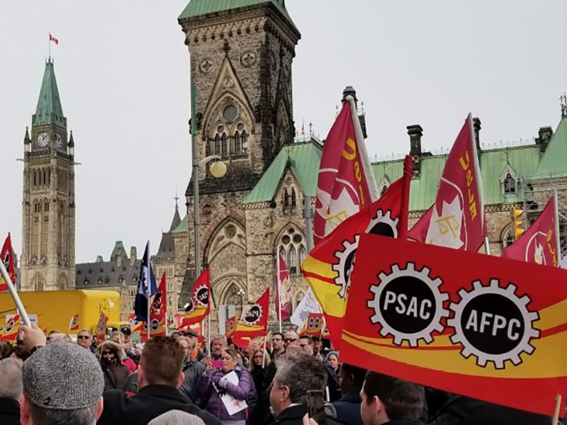 Members of PSAC on a grassy area in Ottawa with the parliamentary buildings in the background. They are holding PSAC flags.