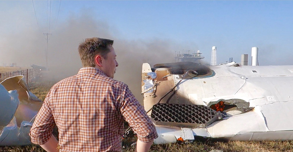 With his hands on his hips, Elon Musk overlooks the wreckage after the failed test launched of an orbital rocket booster in 2014. Smoke fills the air.