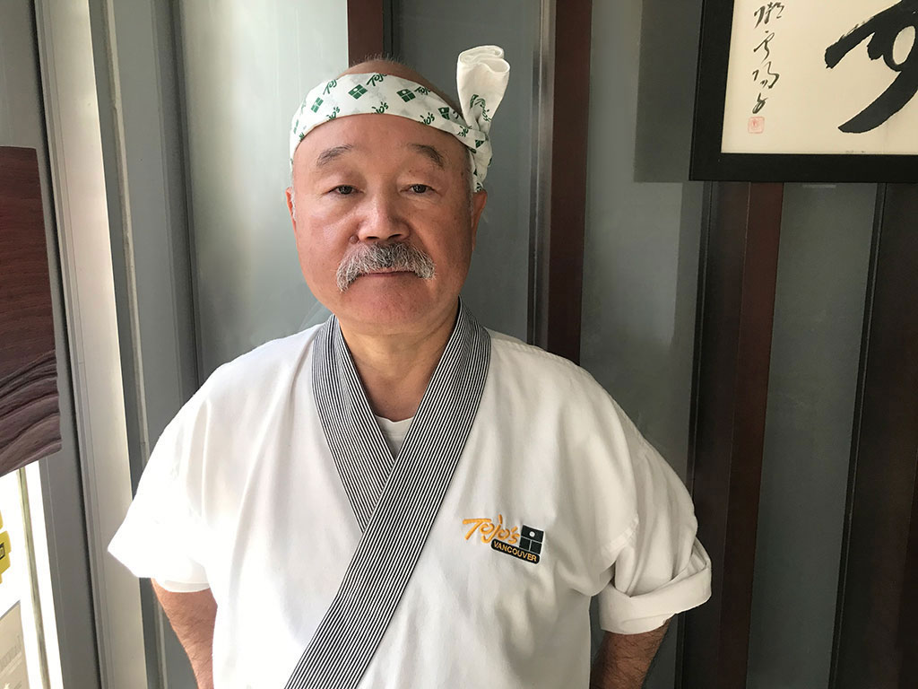 Chef Tojo wears a Tojo’s Vancouver chef’s coat and headband. He has a salt-and-pepper mustache.