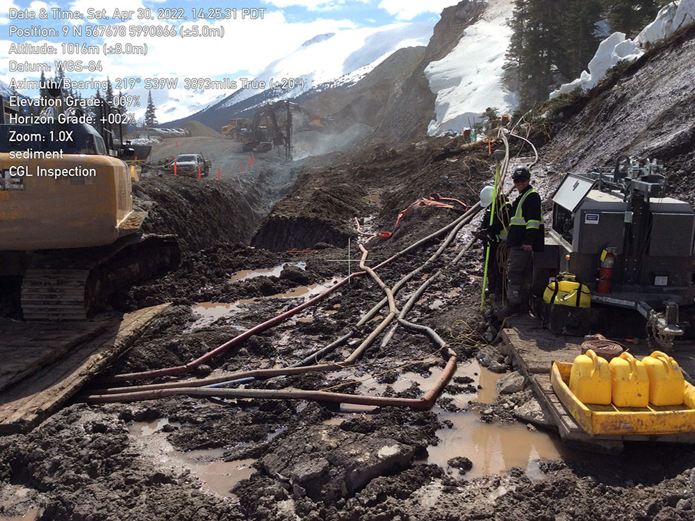 The photo shows heavy equipment, hoses and a long stretch of mud and pools of water along the pipeline construction route. A man stands on one side. Snowbanks overhang the muddy site.