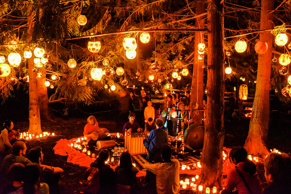 A musical performance by Turtle Bliss Gamelan takes place on the forest floor in Renfrew Ravine at night, with audience members seated nearby on the ground. Handmade paper lanterns decorate the branches of surrounding trees and smaller glass jar lanterns illuminate the forest floor, all part of annual celebrations for the Renfrew Ravine Moon Festival.