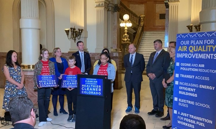 A dozen people, a mix of kids and well-dressed adults, look on as the slight figure of Shaina Oliver speaks from a podium inside the state capitol building, the interior of which includes columns, a stately staircase and glowing, old-fashioned round lamps on stanchions. Blue signs say ‘Healthier Cleaner Colorado’ and a larger blue display lists ‘Our Plan for Air Quality Improvements’ including electric school busses and ‘a just transition for fossil fuel workers.’