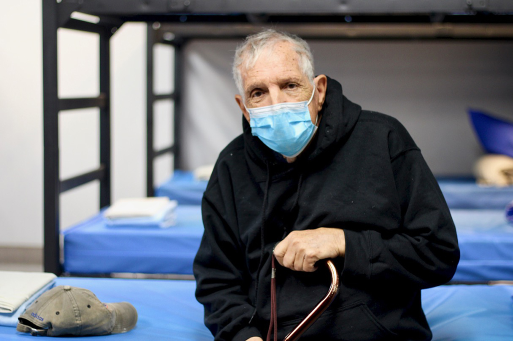 An elderly man sits on a bed in a homeless shelter. Bunk beds can be seen in the background.