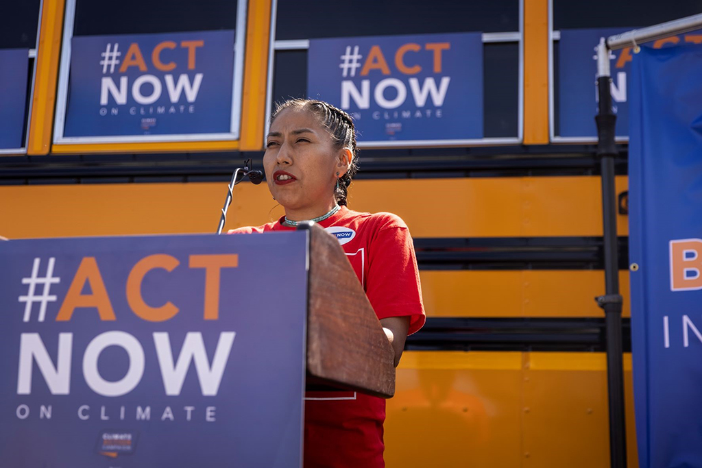 Shaina Oliver, a thin Indigenous woman with braided black hair and wearing a red T-shirt speaks at a podium with signs saying ‘Hashtag Act Now on Climate.’