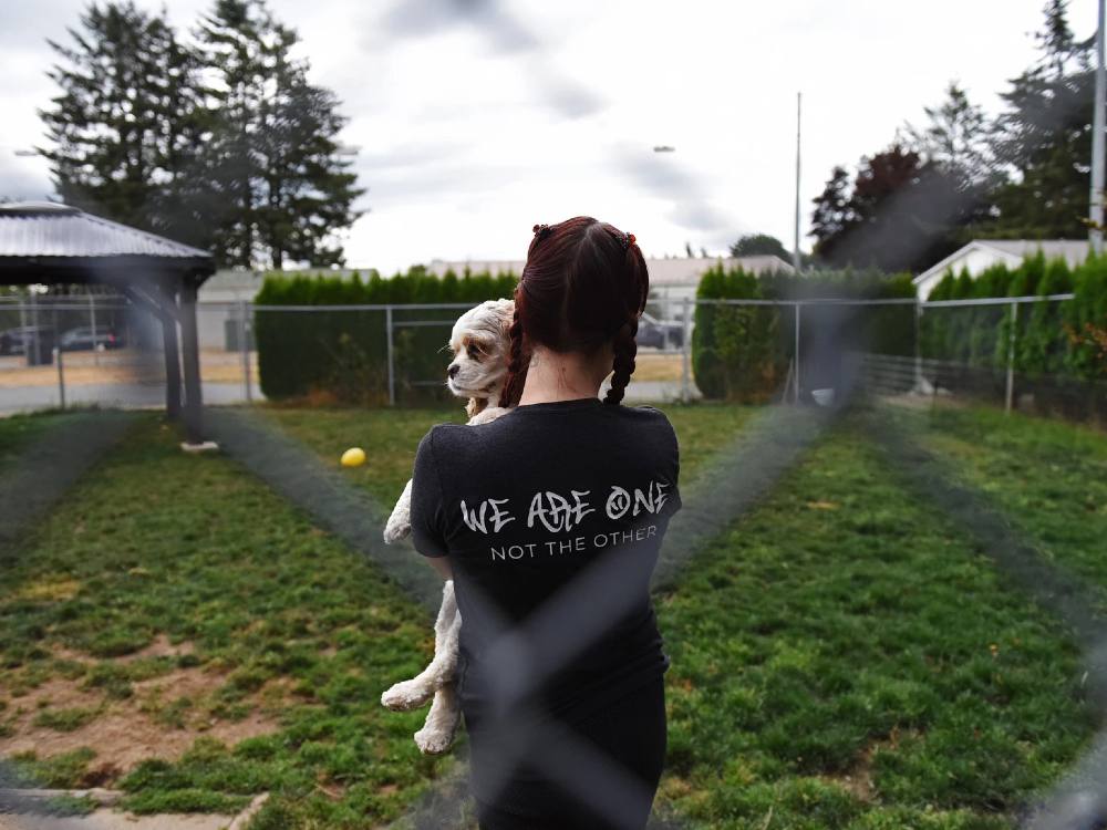 A woman stands outside in a fenced area. She has two braids. She is facing away from the camera and holding a dog in her arms. 