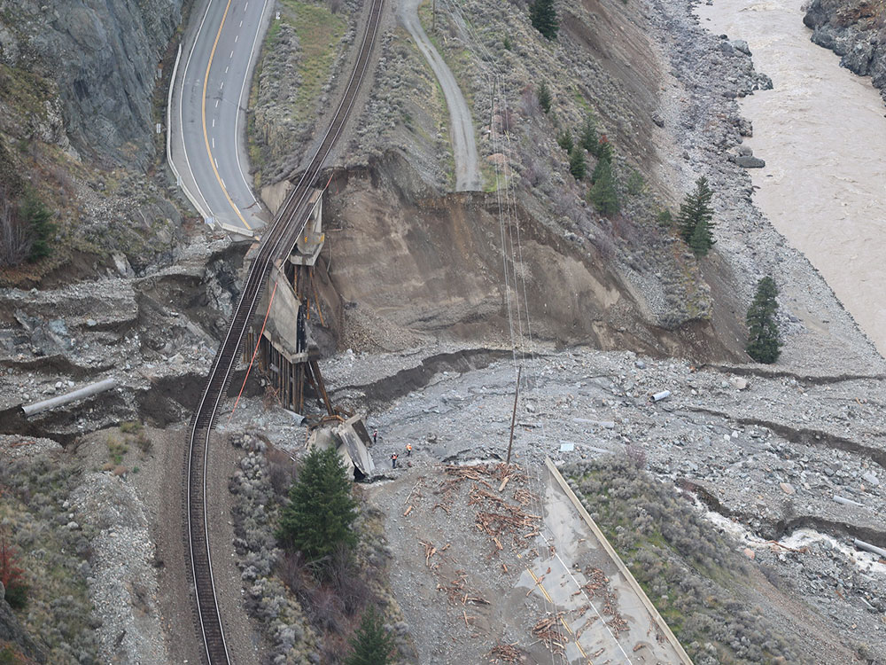An aerial view shows a highway bridge destroyed by floods and landslides and a sagging railroad track beside a river.