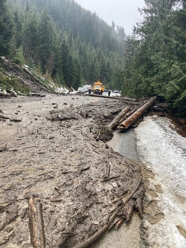 A mudslide covers a road. Beyond the mudslide, cars and people are lined up alongside a yellow emergency vehicle. 