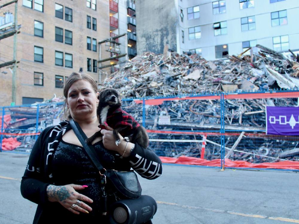 A woman holding a small dog stands on a sidewalk in Vancouver’s Gastown neighbourhood. In the background, a huge pile of rubble from a building that was torn down after a fire sits behind a fence.