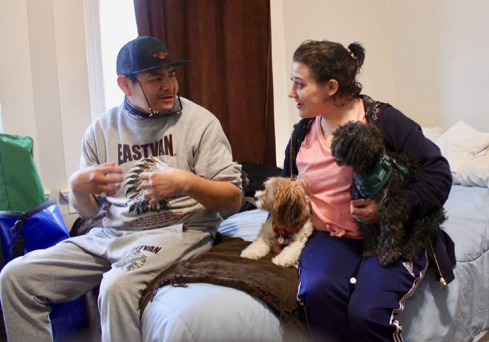 A man and a woman sit on a bed, facing each other in mid-conversation. The woman holds two small dogs, one under each arm.