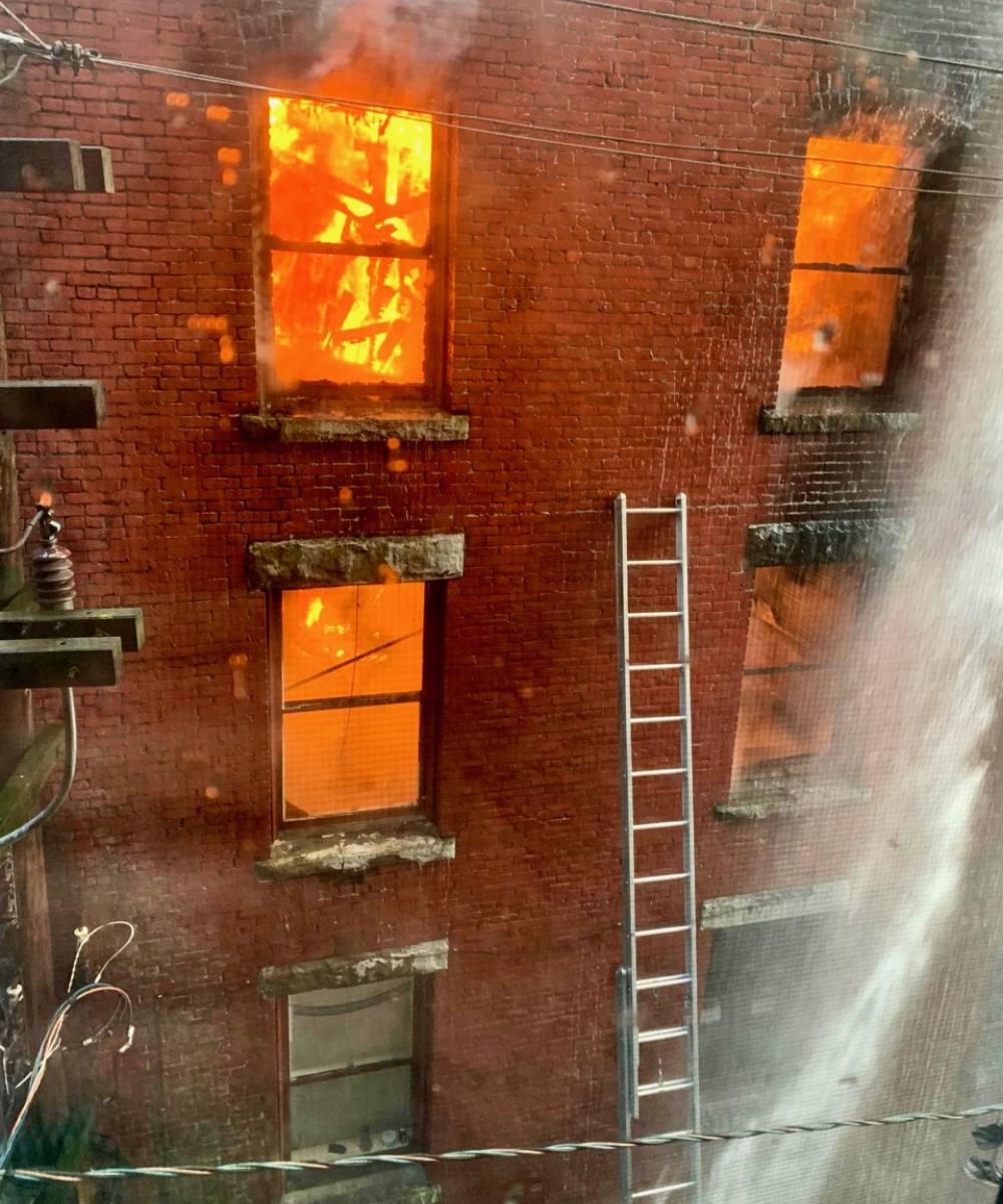 The back of a brick building showing six windows. Four of the windows are filled with yellow and orange flames and smoke is coming out of some of the windows. 