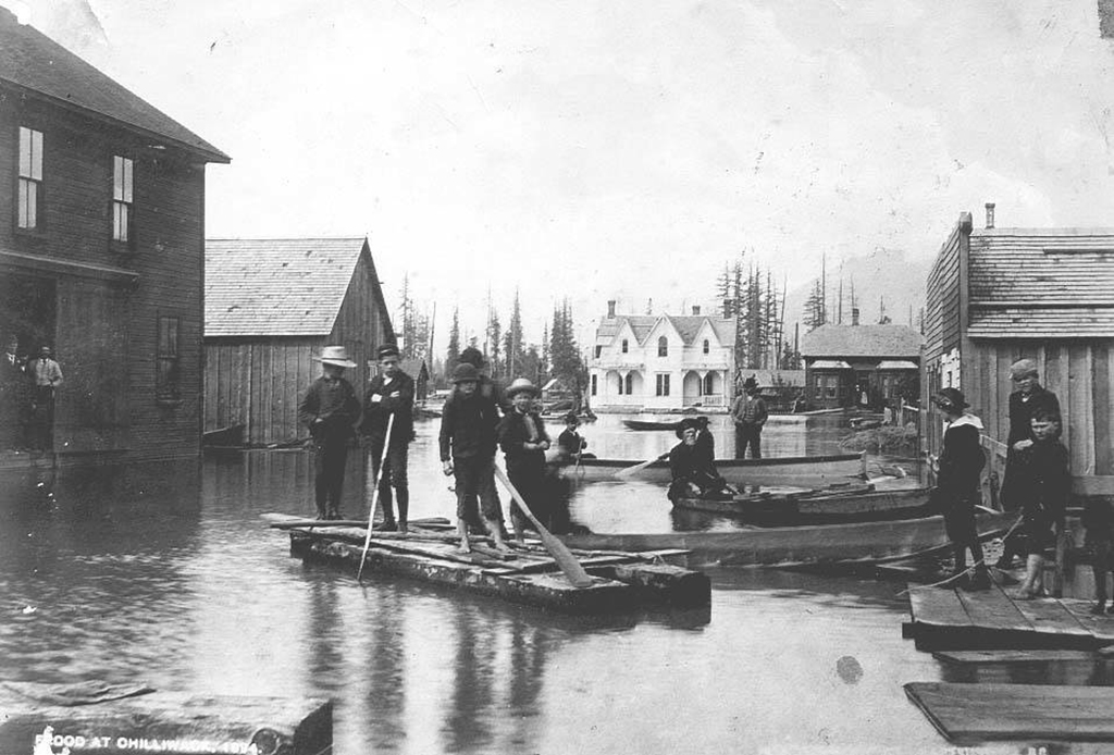 A black and white photo of a flooded neighbourhood. At the centre are half-a-dozen young men on a homemade wooden raft with makeshift oars. There are houses in the background and the floodwaters have risen to the bottom of their doorways.