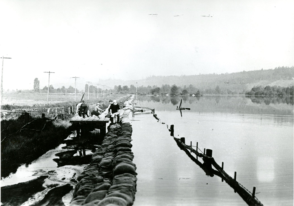 A black and white photo of farmland, with sandbags on the left just barely holding back floodwaters on the right. There are people on an elevated platform adding more sandbags to the barrier.