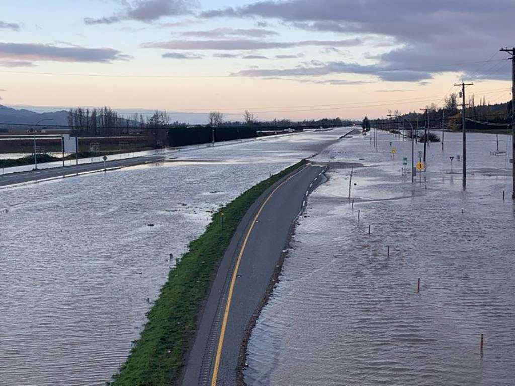 A thin strip of the yellow line that separates traffic stretches off into the distance. It is the only dry land that can be seen in a flooded area. Telephone poles and fence posts stick out of the water, suggesting there is farmland beneath. It’s twilight and the sky is tinged pink and purple.