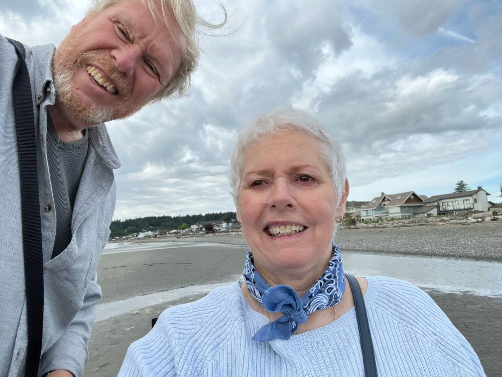 Two people pose for a selfie on the beach near the Boundary Bay neighbourhood of Delta. John Carter, left, is standing holding the camera. He has blonde hair and is wearing a light grey shirt over a grey t-shirt. He is smiling. Beside him, Carolyn Carter is seated in a wheelchair. She has short white hair and is wearing a blue scarf and white sweater. She is smiling.