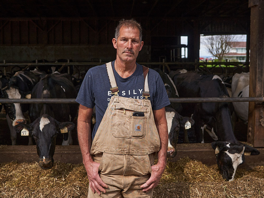 A man in tan overalls and a navy blue t-shirt stands in front of a barn of black and white milking cows at his family’s farm in Pitt Meadows, BC.