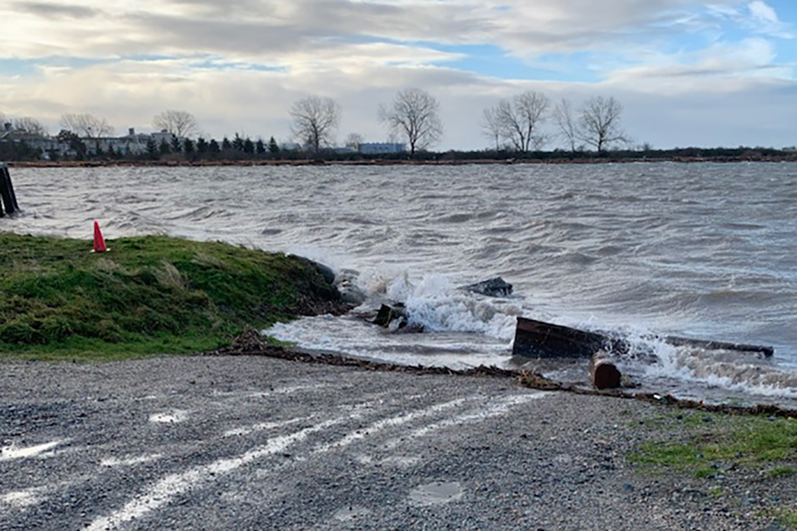 A view of water from a gravelly shore on a cloudy day. Cresting waves spill onto a section of the shore that slopes down to the water, where a ramp used to be. There is an orange cone at the edge of the shore where it meets the river.