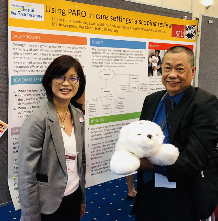 Two people stand in front of a poster board with the headline ‘Using Paro in care settings: a scoping review.’ They are IDEA Lab founder Lillian Hung, left, and ‘patient-partner’ Mario Gregorio, right. They both have black hair. Lillian is wearing glasses and a grey blazer. William has shorter hair and is wearing a black suit jacket over a blue shirt. He is holding what looks like a white furry stuffed animal called the Paro Robot.