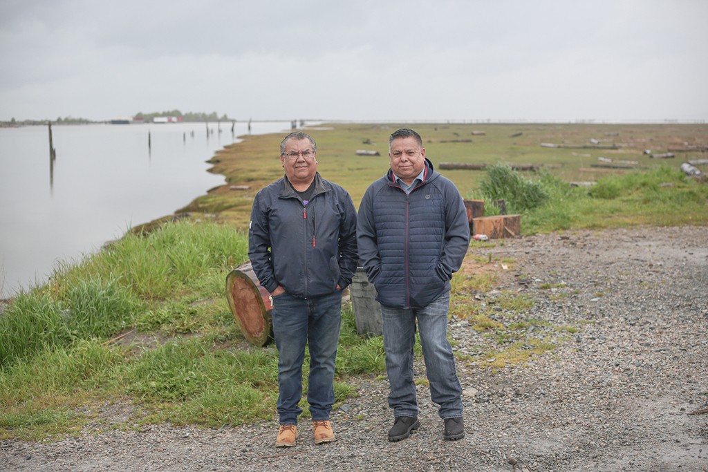 Two brothers, 59 and 60, stand on gravel by the edge of the Fraser River. This is where the river meets the sea and there is open water in the background. Along the shore of the river is a grassy marsh, visible because the tide is out.