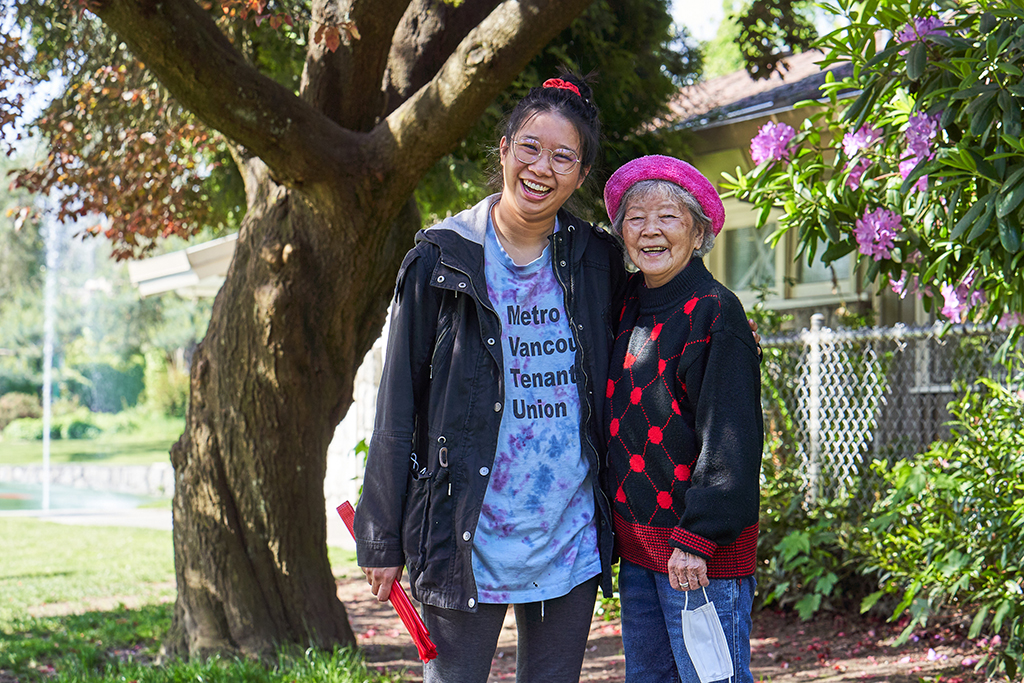 Two people stand together, smiling at the camera. Beverly Ho, left, is wearing a blue T-shirt and black jacket. Xing Jun Ma, right, is wearing a pink cap and a black and red patterned sweater.