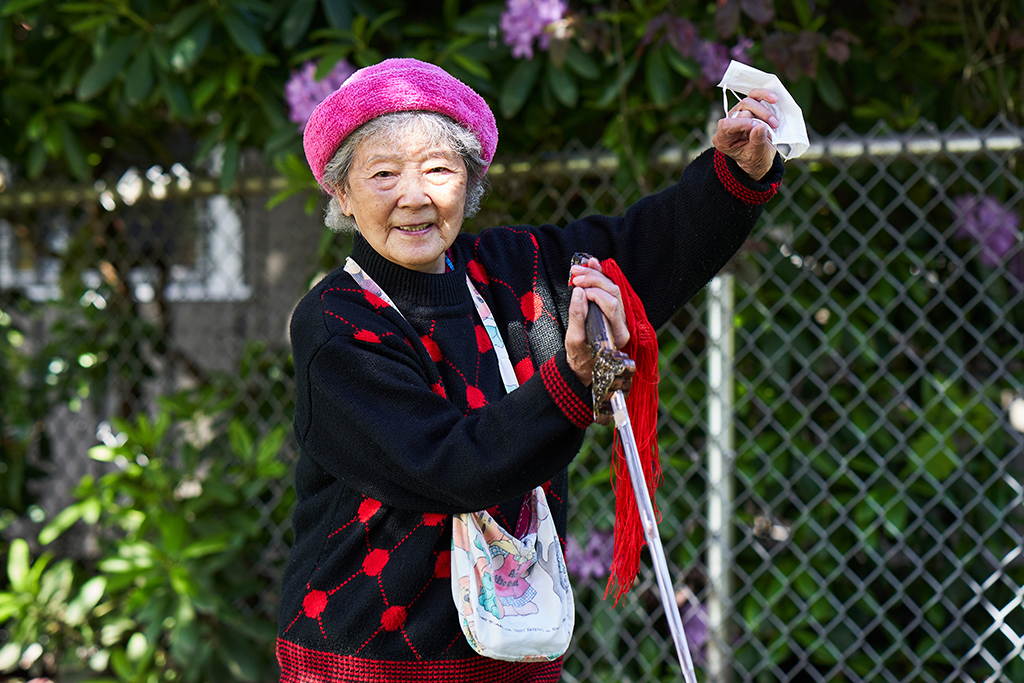 A 90-year-old woman, Xing Jun Ma, poses holding a tai chi sword and a medical mask aloft. She is wearing a fuchsia cap and a black sweater with bright red polka dots.