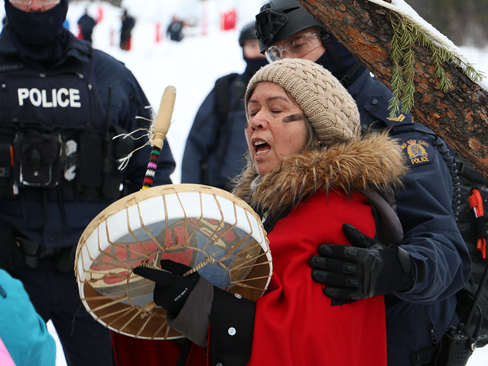 In a snowy setting, a woman bangs a hand-drum as police take her into custody.