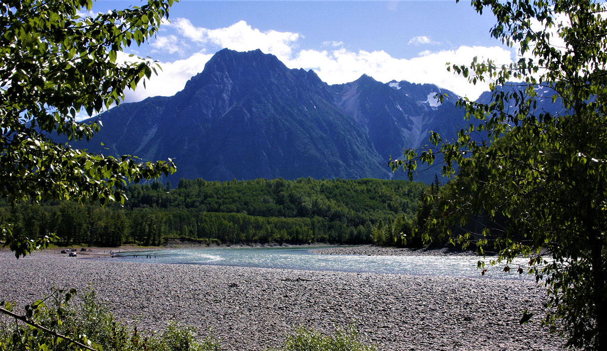 A blue mountain with some snow rises behind forest and in the foreground a river with a broad, flat gravelly bank. 