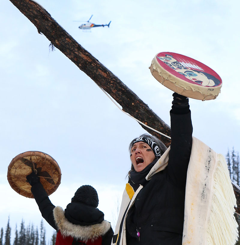 Freda Huson hitting a drum in her hand and looking up and yelling, a fellow protester nearby and a helicopter hovering overhead.