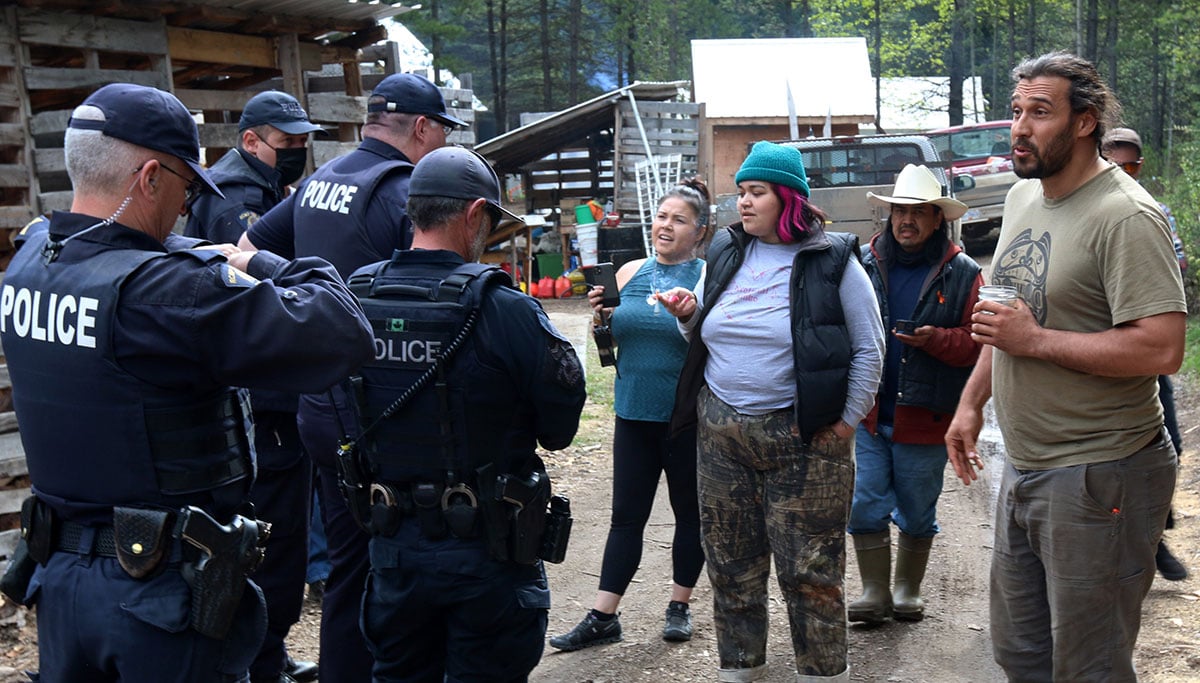 A handful of police face a handful of animated people in an encampment in a forest clearing. 