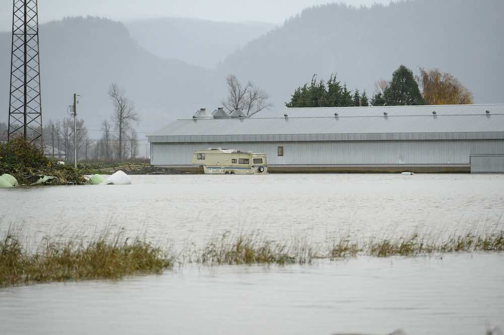 In the foreground, floodwaters cover farmland. In the near distance is a flat, white, light blue building with a beige camper van in front of it. Misty mountains are visible in the background.