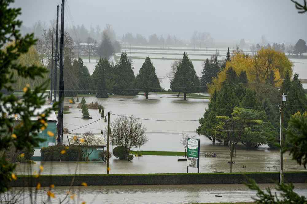 A grey, rainy landscape is dotted with evergreen trees. In the foreground is a green sign for the Fraserglen Golf & Training Centre. The golf course and driving range is covered in water.