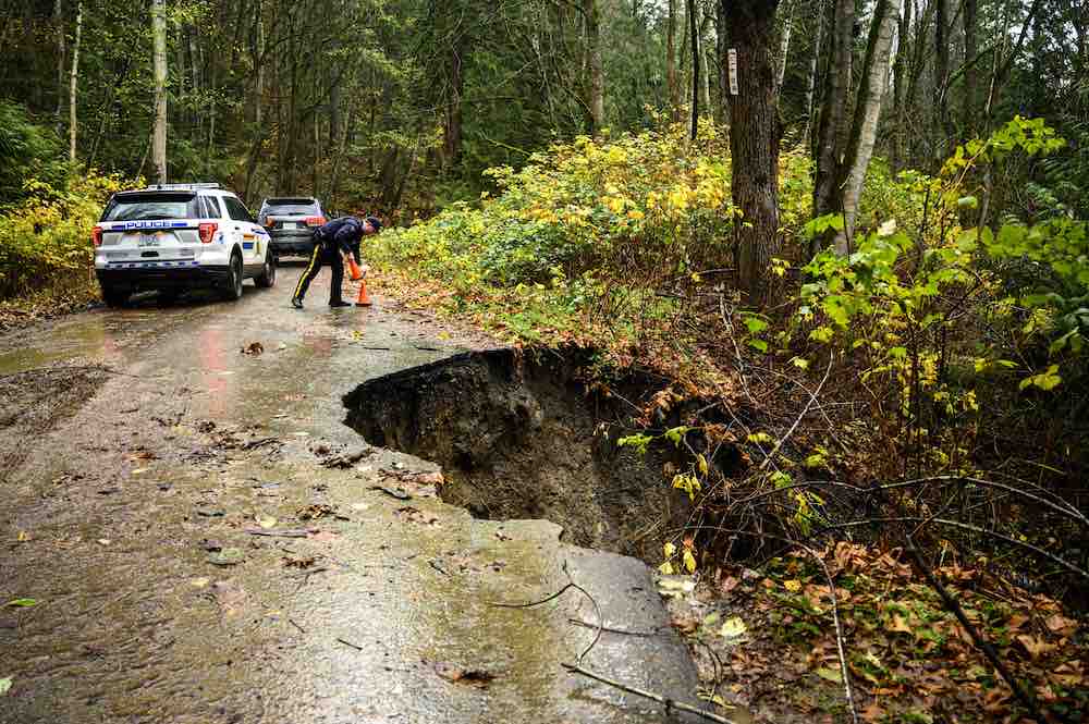 A large hole is visible on the side of a forest road. A police offer bends to place a orange pylon on the road. They are standing behind two sports utility vehicles to the left of the frame.