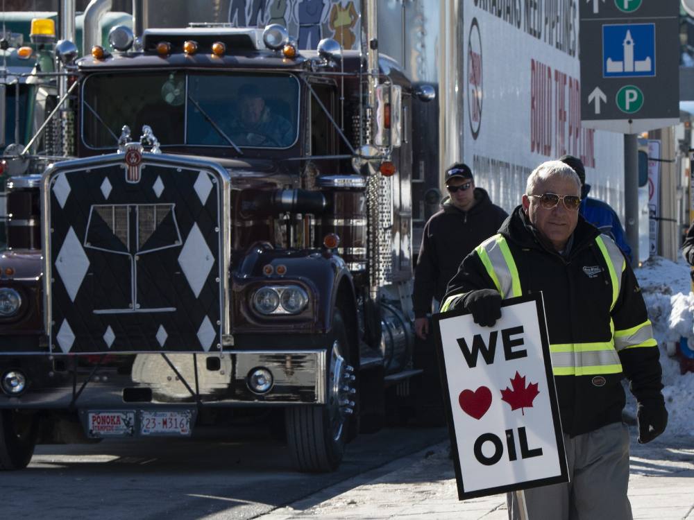 A man with grey hair and a high-vis vest stands in front of a large truck, holding a sign saying "we love Canadian oil."  
