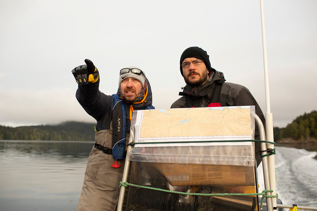 Two warmly dressed men squint against the wind as they consult a map on a boat. Pacific Salmon Foundation fish pathologist Emiliano Di Cicco is on the left, pointing at something in the distance over the photographer's shoulder.
