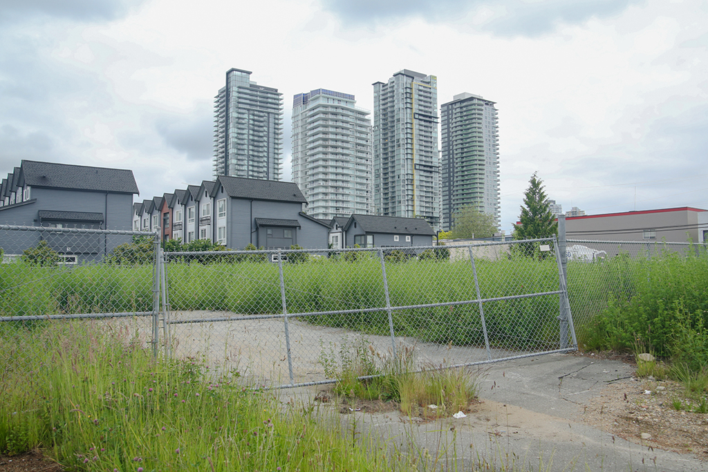An empty lot with a tilted chain-link gate that looks like it’s about to fall off from a chain-link fence. The lot is covered with tall grass. In the background are three rows of townhouses with pitched roofs, and behind them are four sleek, glassy condo towers against a cloudy sky.