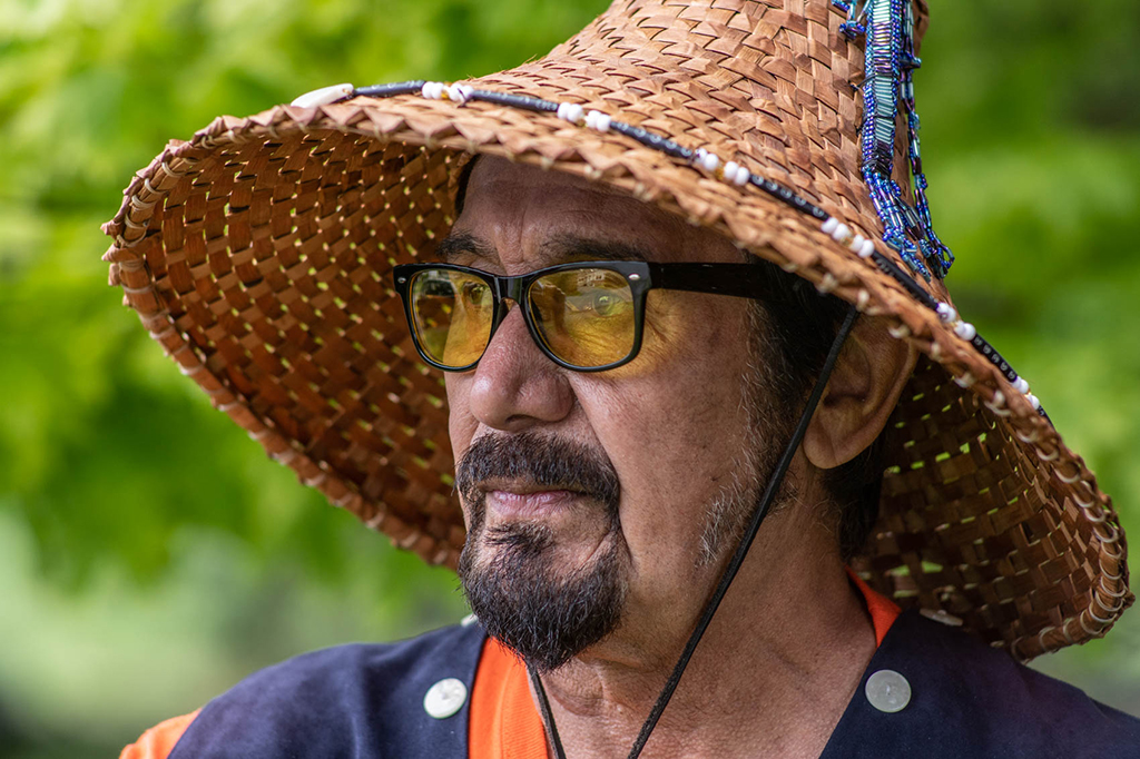 Hereditary Chief Tsahavkuse wears a cedar bark hat and a button blanket vest. His wears his facial hair in a tidy goatee. His black frame glasses have a yellow tinted frame.