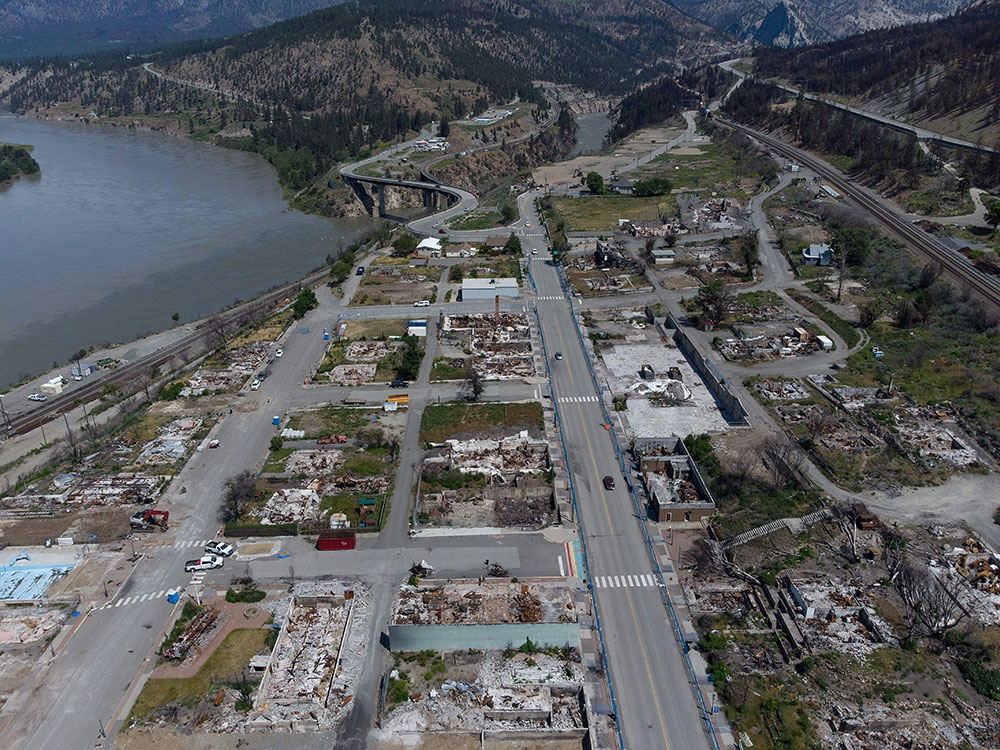 An aerial photo shows about 16 blocks of Lytton after the fire. The roads are intact but no buildings are standing and most are rubble. The Fraser River is on the left and railway tracks run along the other side of town.