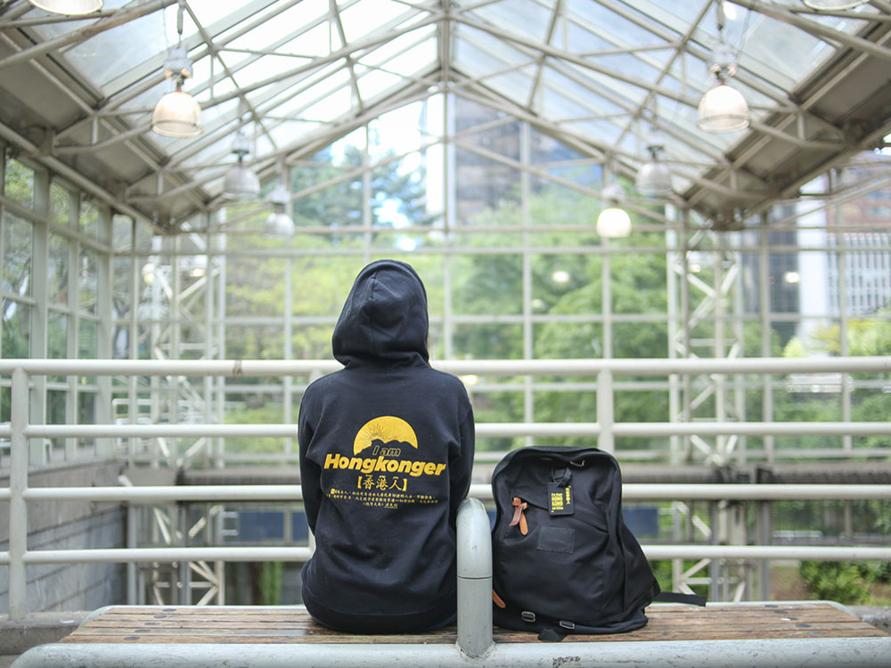 A woman sits on a bench at a transit station with her back to the camera. She’s wearing a black hoodie, with yellow text on the back that says ‘I am Hongkonger’ in English and ‘Hongkonger’ in Chinese characters. She has a black backpack beside her with a tag that says, ‘I’m from Hong Kong, not China.’