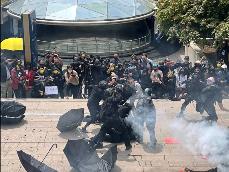 A crowd watches the re-enactment of 2019’s protests in Hong Kong in a downtown plaza. There are umbrellas scattered on the ground as actors recreate a chaotic scene of police beating protesters with batons.