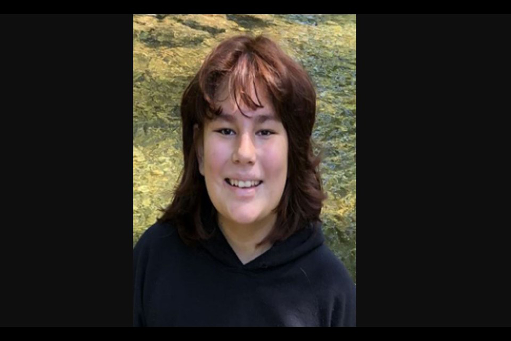 A young person with auburn shoulder-length hair, wearing a black hoodie, smiles at the camera.