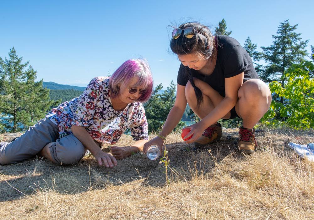 One person lies down, and one crouches, on dry grass. The person on the right is pouring water onto the ground from a glass jar.