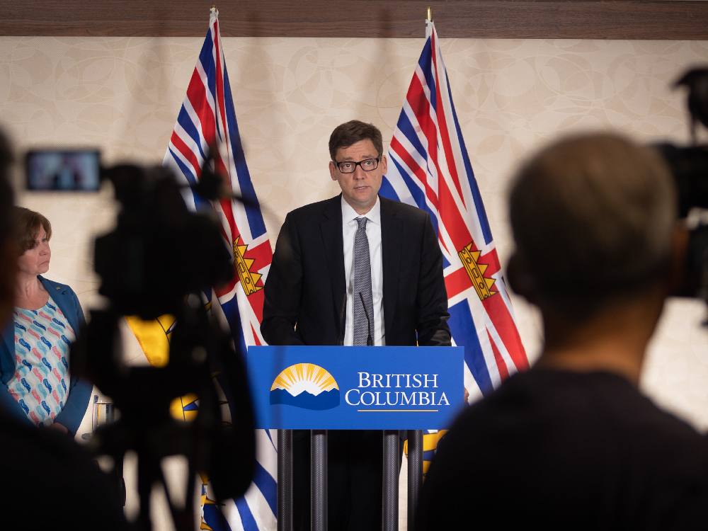 BC's Attorney General David Eby stands in front of a podium flanked by two BC flags. 