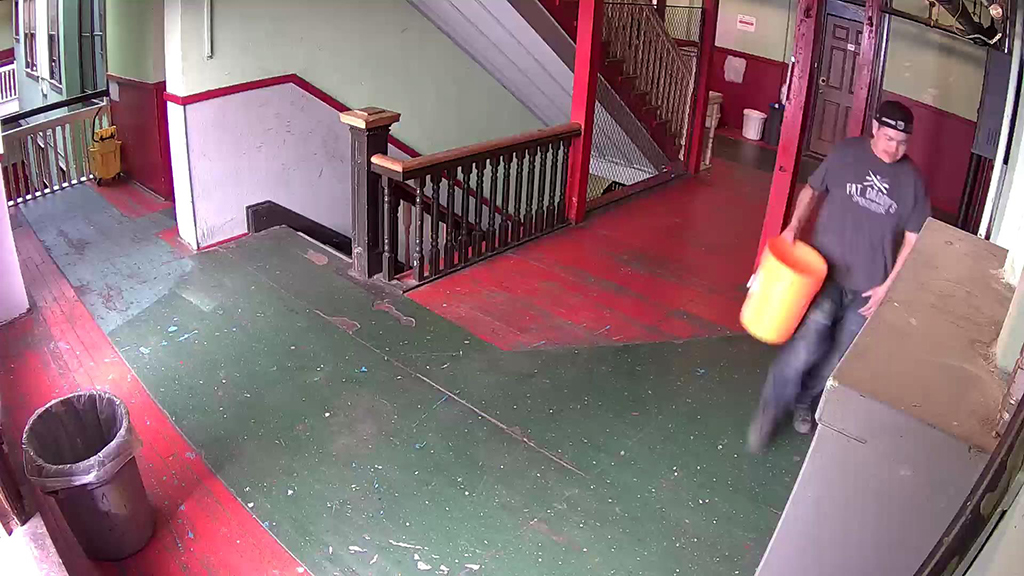 A man wearing a backward ball cap, T-shirt and jeans stands in a hallway of an old building with a worn green and red wood floor. He holds a yellow plastic bucket in one hand.