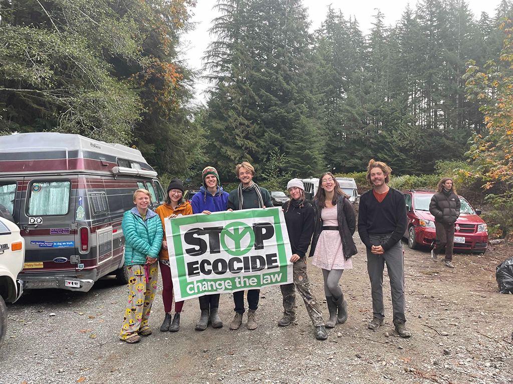 Seven young people stand next to each other, smiling for the camera. They are standing in a parking area and behind them are tall trees. They are protesters part of the Fairy Creek blockade in summer 2021. Jamie Hunter is wearing a bright blue jacket and is third from the left. He is holding a green and white sign that reads “Stop ecocide, change the law.”