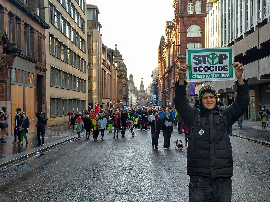 Jamie Hunter is wearing a green and red toque, quilted black jacket and jeans. He is holding a green and white sign over his head that reads “Stop Ecocide, change the law.” Behind him, people march in the streets of Glasgow, Scotland as the sun sets. They are part of the COP26 conference in 2021.