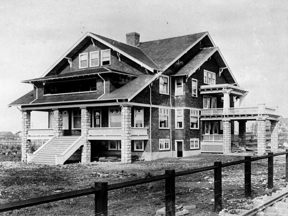 A black and white archival photo of a relatively large building covered in cedar shakes. The front of the building has two covered porches. There is a second entrance at the side of the building.