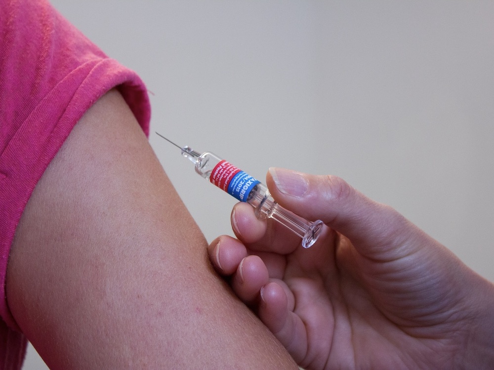 A syringe is poised to inject into the upper arm of a person wearing a pink T-shirt.