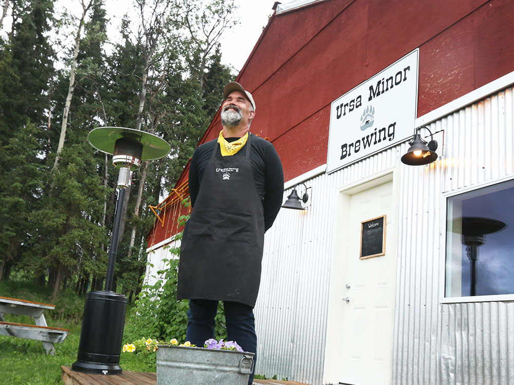 A grinning, bearded man wearing a ball cap, yellow bandana around his neck and black pants, shirt and apron, stands on a deck in front of a building. A sign over the door says 'Ursa Minor Brewing.'