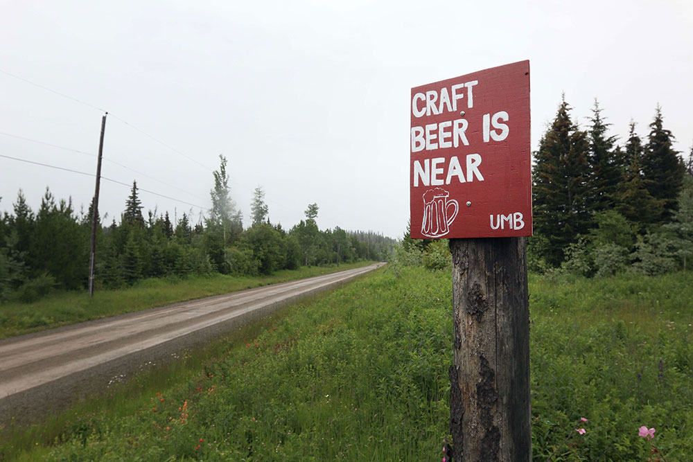 A dirt road stretches into the distance, with trees on either side. On a pole, there’s a red sign with white letters saying ‘Craft beer is near.’